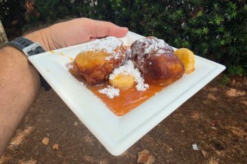 Hand holding a square plate with two fried dough balls dusted with powdered sugar, resting in caramel sauce with a lemon wedge, outdoors.