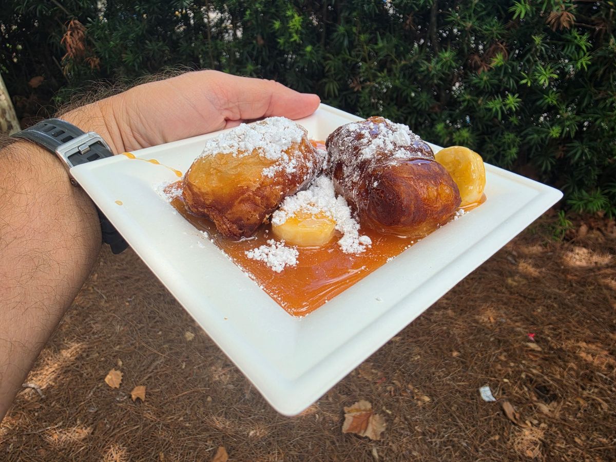 Hand holding a square plate with two fried dough balls dusted with powdered sugar, resting in caramel sauce with a lemon wedge, outdoors.