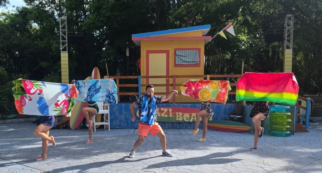 Group of five people dancing with colorful towels over their shoulders on a sunny outdoor stage with a small yellow hut behind them for Summer Nights 2026