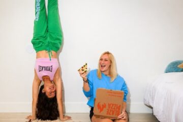Two women in a bright room: one does a handstand in green pants and a pink top, the other sits on the floor in a blue sweater, laughing with pizza and a box nearby.