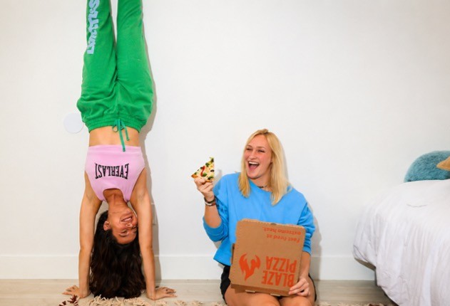 Two women in a bright room: one does a handstand in green pants and a pink top, the other sits on the floor in a blue sweater, laughing with pizza and a box nearby.
