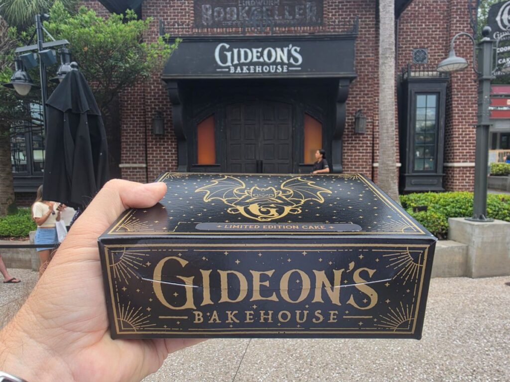 Person holds a black-and-gold Gideon's Bakehouse cake box in front of the bakery storefront sign.