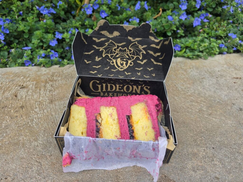 Opened Gideon's Bakehouse box holding the Blueberry Lemonade Cake showing a pink-frosted cake slice with yellow sponge layers on parchment, outdoors among blue flowers.