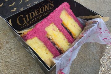 Pink and yellow layered Blueberry Lemonade cake slice in a Gideon's Bakehouse box on a concrete surface, with parchment paper.