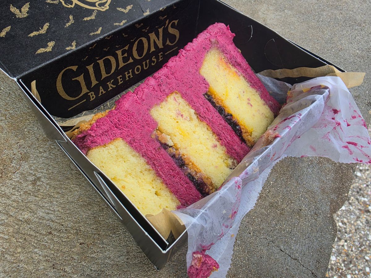 Pink and yellow layered Blueberry Lemonade cake slice in a Gideon's Bakehouse box on a concrete surface, with parchment paper.