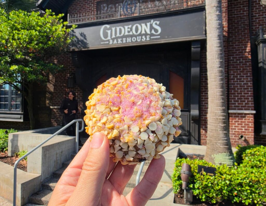Hand holding a round confection coated in white chips and pink sugar outside Gideon's Bakehouse storefront.