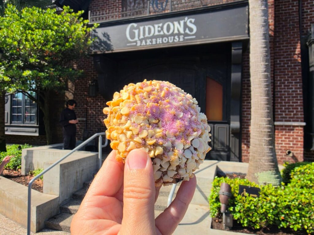 Close-up of a round Blueberry Lemon Cookie held in a hand, with Gideon's Bakehouse storefront in the background.