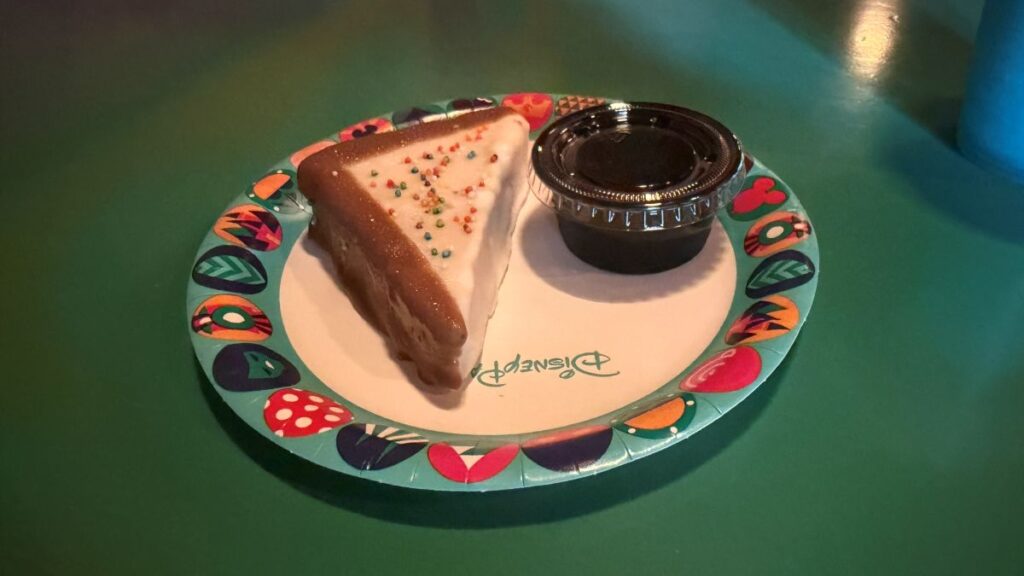 Triangular slice of vanilla cake with white frosting and colorful sprinkles on a festive paper plate, next to a small black sauce cup on a green table for Pizzafari Bluey menu review