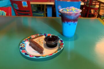 Triangular slice of chocolate cake with rainbow sprinkles on a colorful plate, beside a small dipping cup on a turquoise table; a blue layered drink topped with sprinkles is nearby for Pizzafari Bluey menu review