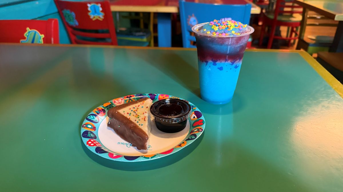 Triangular slice of chocolate cake with rainbow sprinkles on a colorful plate, beside a small dipping cup on a turquoise table; a blue layered drink topped with sprinkles is nearby for Pizzafari Bluey menu review