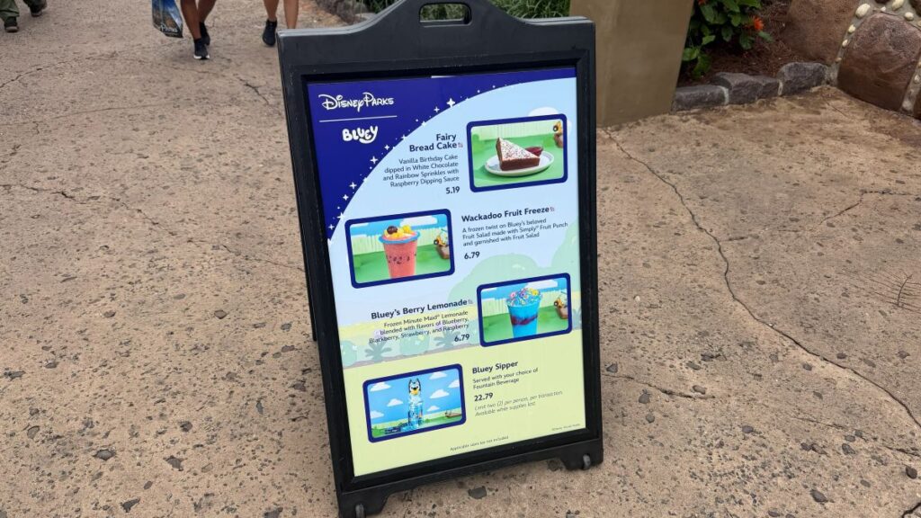 Disney Parks menu board titled 'Bluey' with images and descriptions of snacks: Bread Cake, Wackadoo Fruit Freeze, Berry Lemonade, and Bluey Sipper items on a colorful sign.