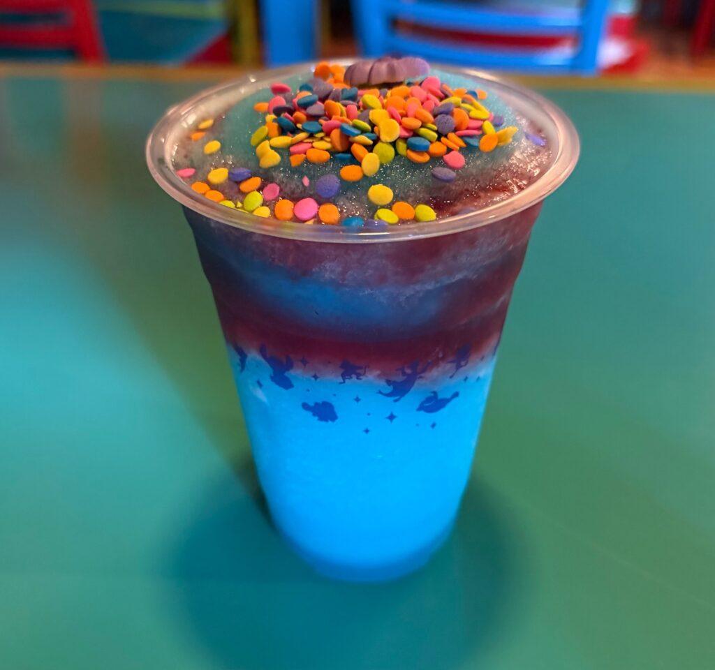 Close-up of a blue gradient drink in a cup topped with colorful rainbow sprinkles on a clear lid, outdoors by a teal surface for Pizzafari Bluey menu review