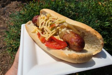Bratwurst in a bun with sautéed peppers, onions, and shredded cheese on a white plate outdoors at SeaWorld Orlando