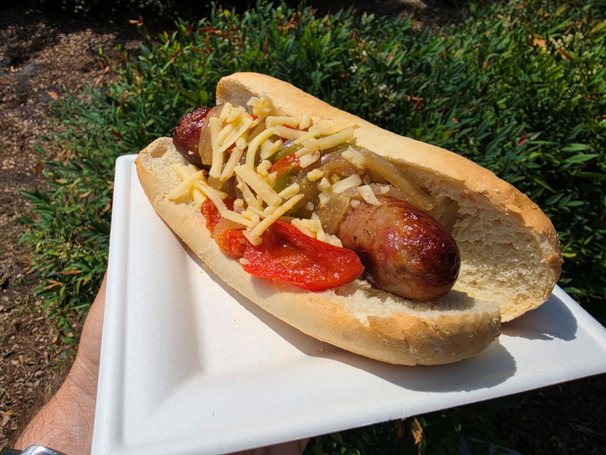 Bratwurst in a bun with sautéed peppers, onions, and shredded cheese on a white plate outdoors at SeaWorld Orlando