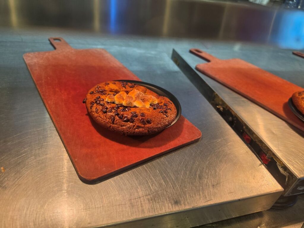 Round Café 4 S'mores Cookie topped with marshmallows and chocolate chips on a red cutting board atop a stainless-steel counter in a bakery kitchen