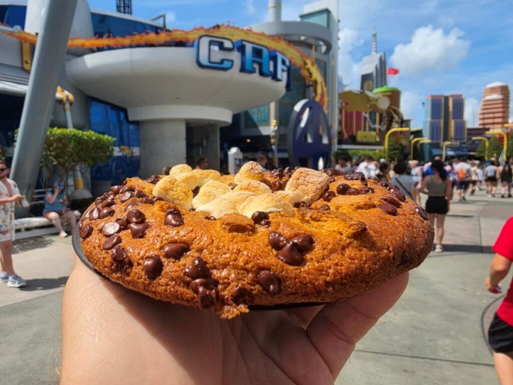 Hand holding a oversized cookie topped with chocolate chips and mini marshmallows at a busy outdoor theme park.