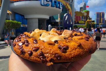 Close-up of a hand holding a large Café 4 S'mores Cookie with mini marshmallows, against a bustling amusement park backdrop.