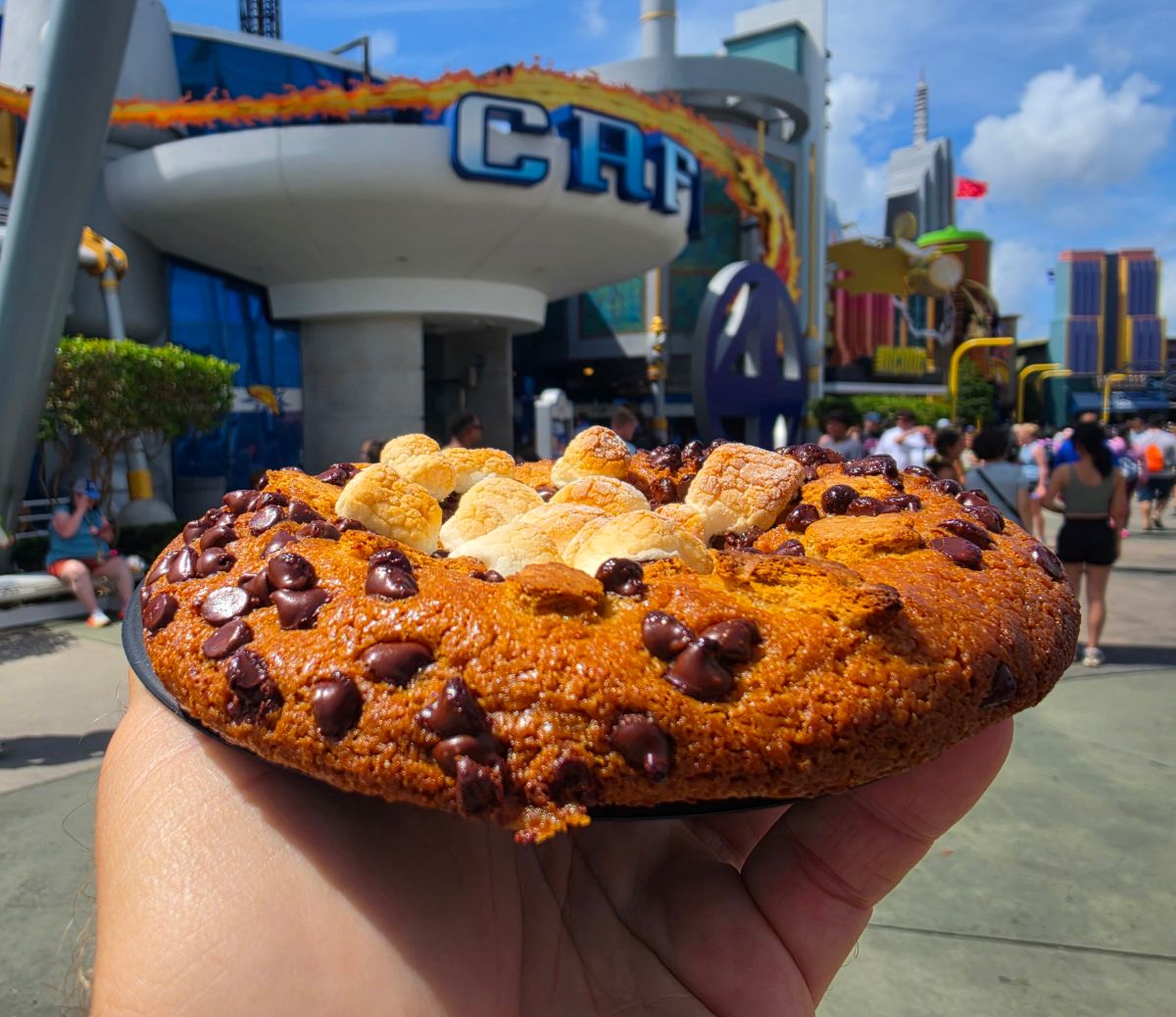 Close-up of a hand holding a large Café 4 S'mores Cookie with mini marshmallows, against a bustling amusement park backdrop.