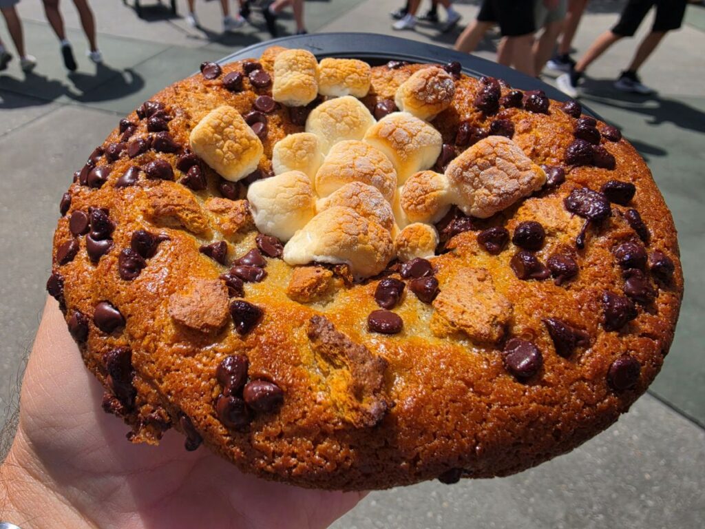 Hand holding a large Café 4 S'mores Cookie topped with marshmallows and chocolate chips outdoors.