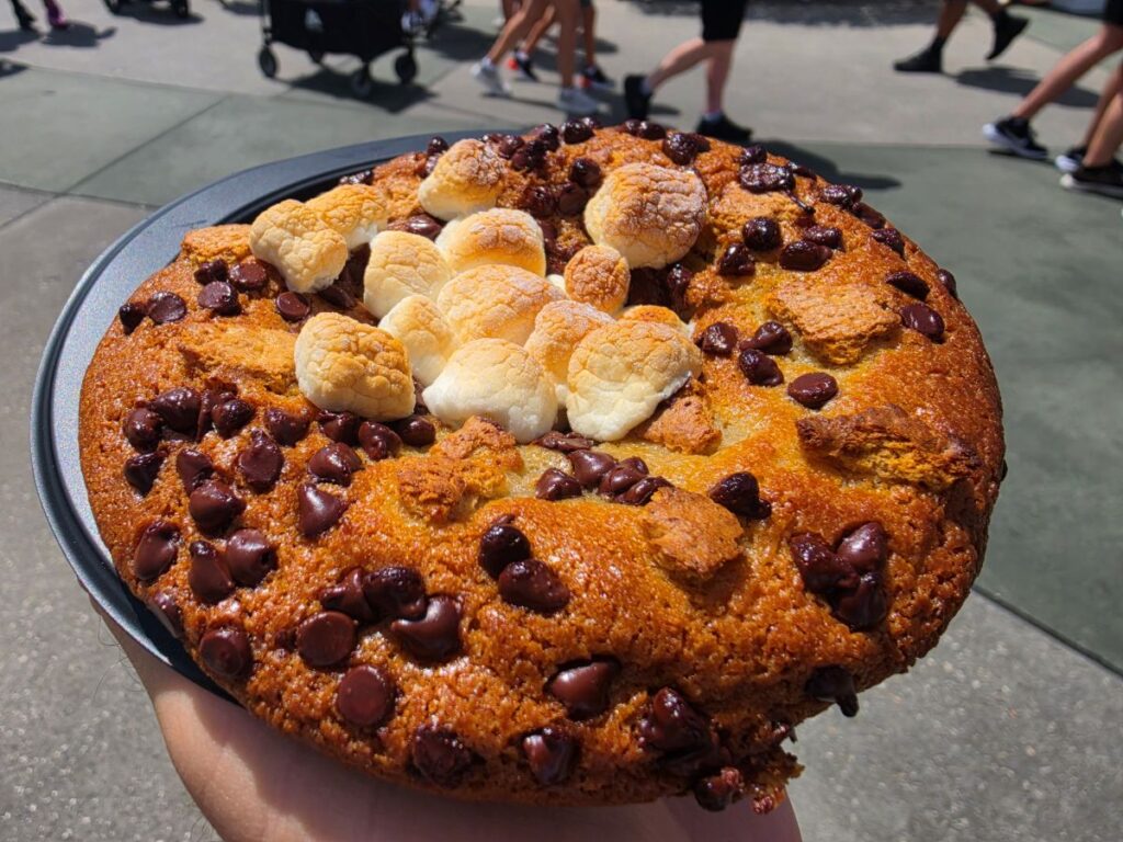 Large round cookie cake topped with mini marshmallows and chocolate chips being held outdoors. People walking in the background.