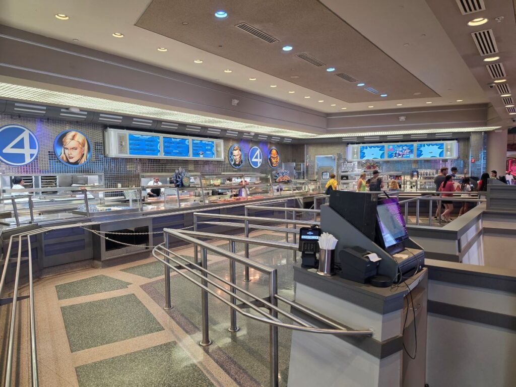 Bright, futuristic food court with service counters, glass display cases, and customers lined up along stainless railings.