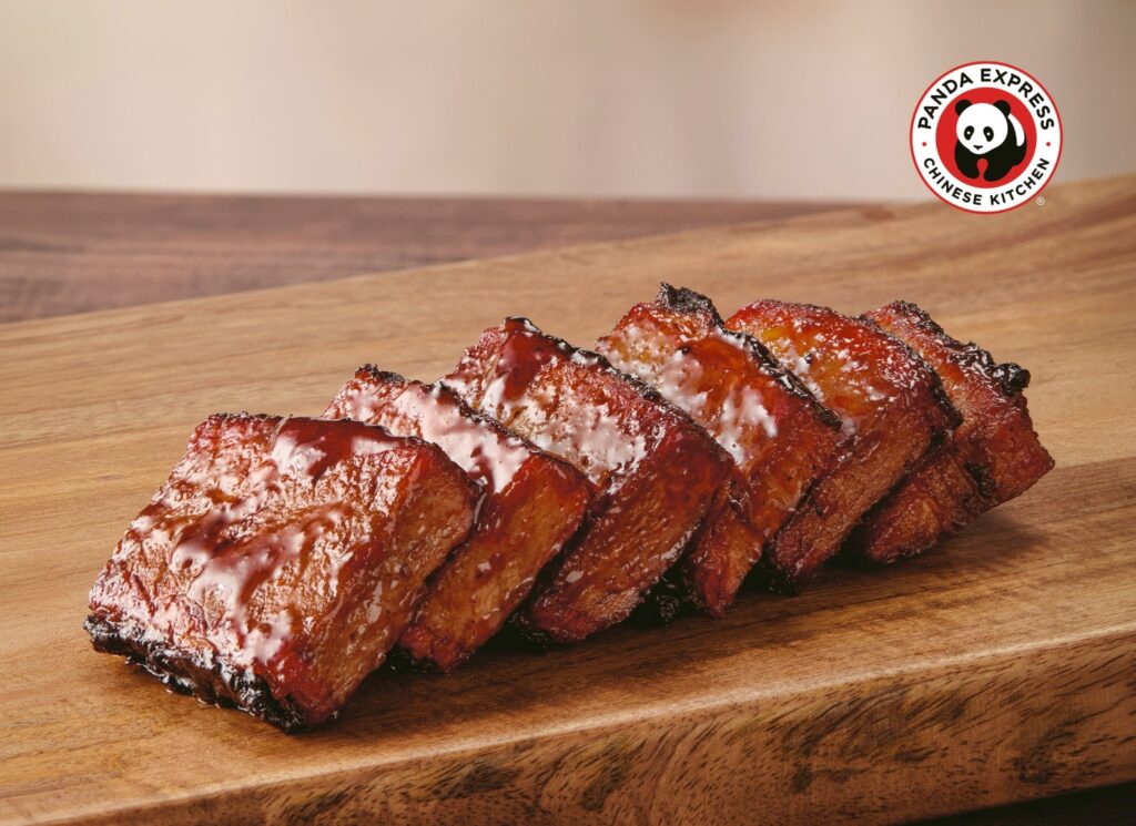Glazed pork ribs stacked on a wooden cutting board with a Panda Express logo in the background (right).
