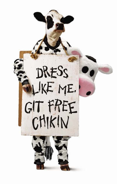 Person in a cow costume holds a sign reading 'DRESS LIKE ME, GET FREE CHIKIN' with a plush cow peeking from behind the sign fro Cow Appreciation Day returns