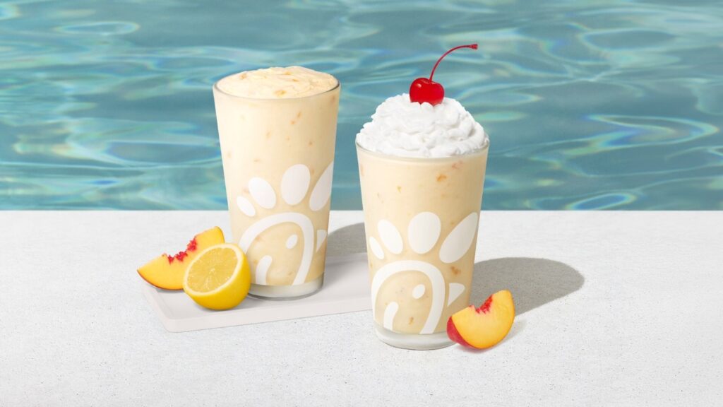 Two peach milkshakes with whipped cream and a cherry on top, poolside with peach slices nearby
