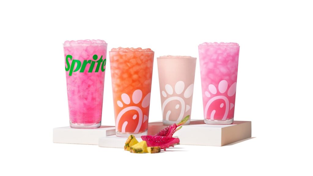 Four colorful iced drinks in tall cups arranged on white blocks, with pineapple and dragon fruit slices in front.