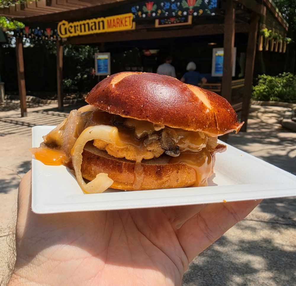 A juicy chicken schnitzel on a glossy pretzel bun with onions and melted cheese, held on a white plate outdoors near a German Market sign at Seven Seas Food festival