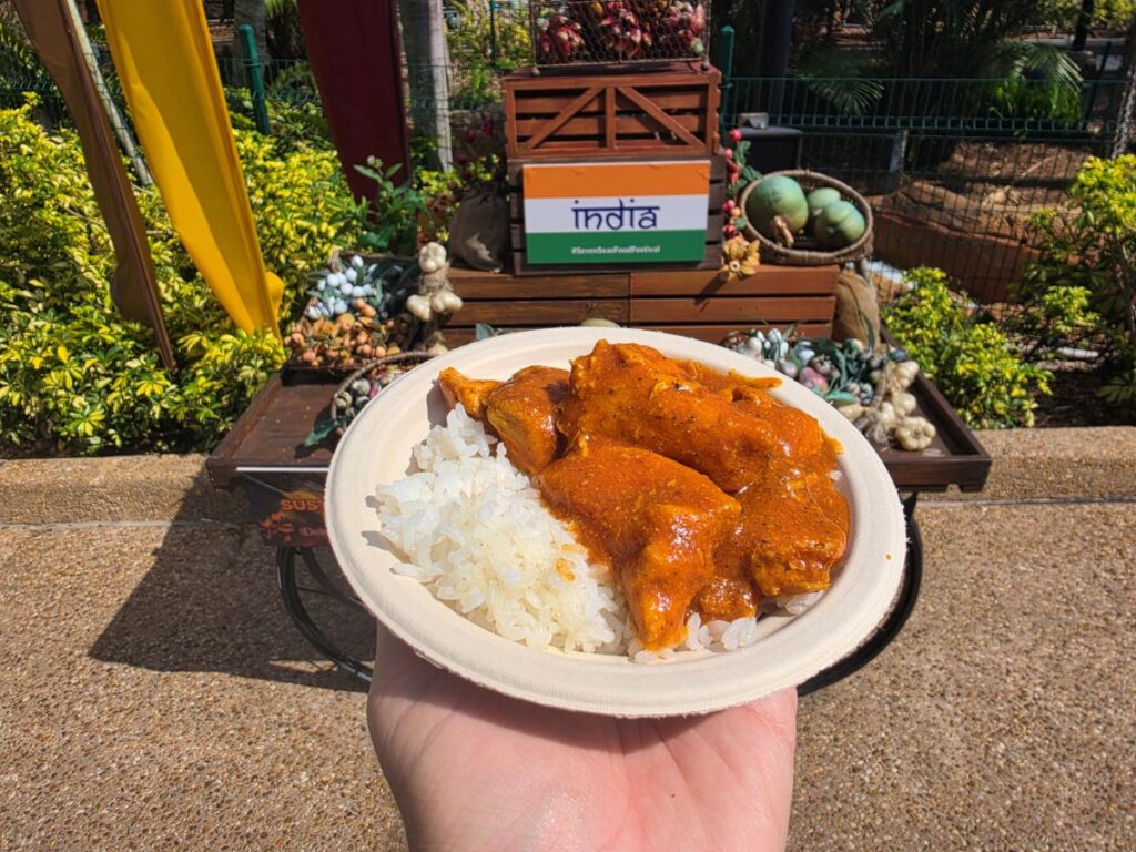 Plate of Chicken Tikka masala with white rice held outdoors at a food festival, with an India sign in the background.