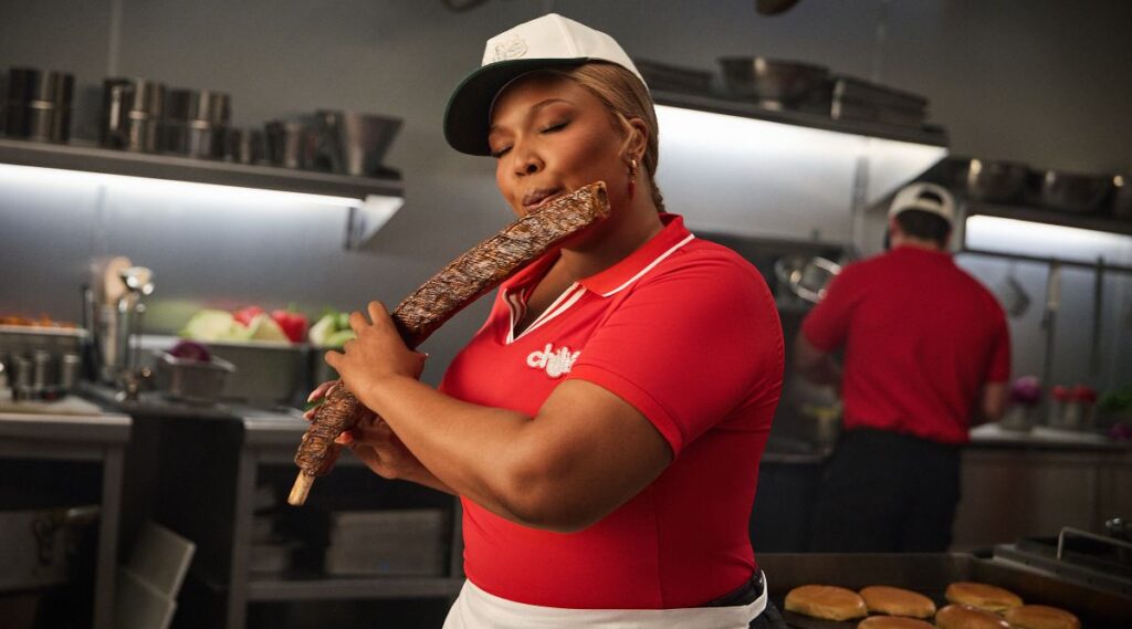 Female worker in a red polo and white cap pretending to play music with a side of ribs.in Lizzo Chili's Ribs Jingle commercial kitchen.