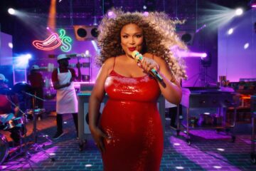Singer in a shiny red dress sings into a microphone on a nightclub stage, colorful lights and a band behind her for Lizzo Chili's Ribs Jingle