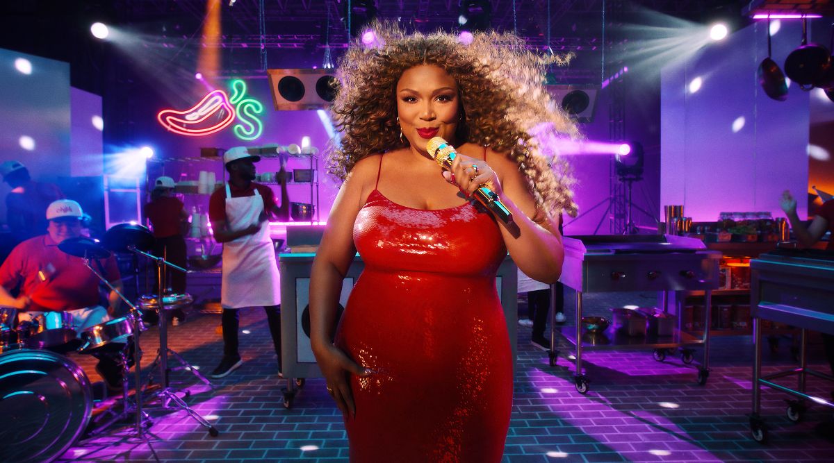 Singer in a shiny red dress sings into a microphone on a nightclub stage, colorful lights and a band behind her for Lizzo Chili's Ribs Jingle