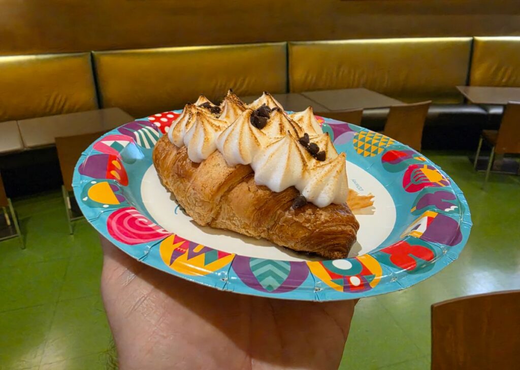 Pastry loaf with toasted cream dollops on top, served on a bright, patterned disposable plate and held in a hand indoors.