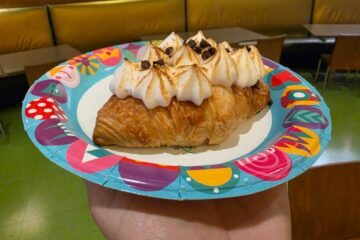 Chocolate Marshmallow Hazelnut Croissant with whipped cream peaks and chocolate chips on a bright, decorative plate in a cafe setting.