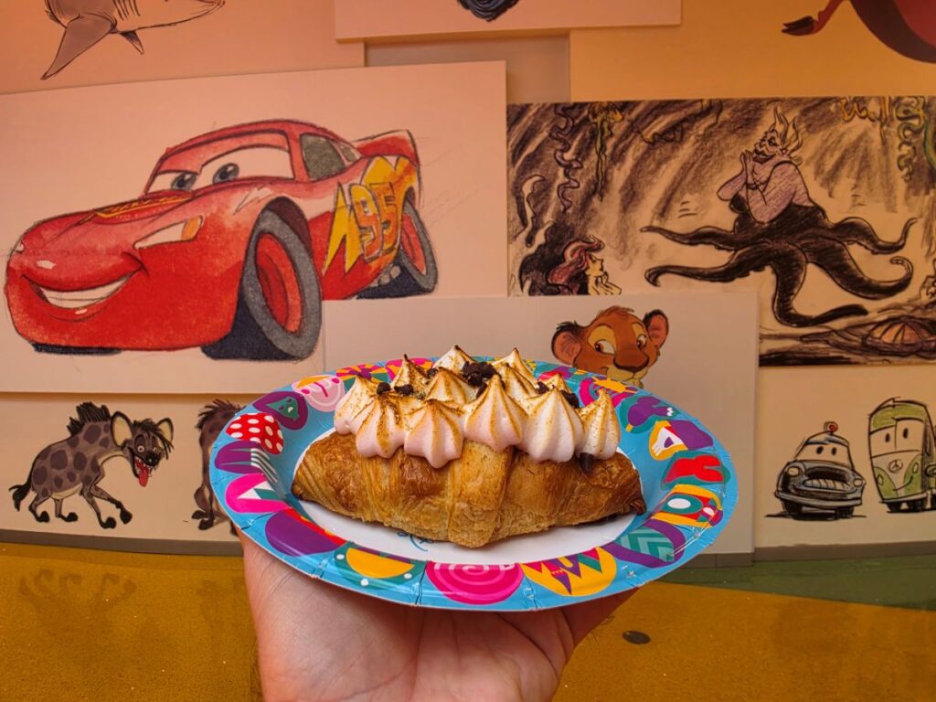 Croissant with piped cream and toasted peaks on a colorful disposable plate held up in front of wall art with cartoon drawings.