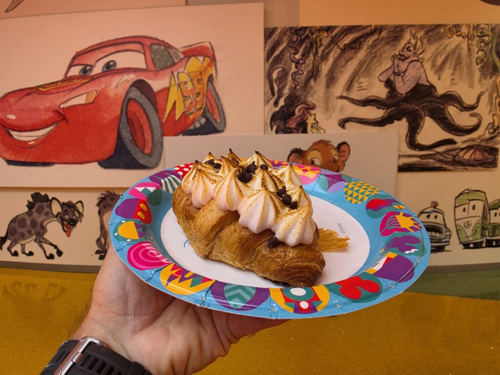 Hand holding a colorful paper plate with a Chocolate Marshmallow Hazelnut Croissant and chocolate chips in front of cartoon wall art, including Lightning McQueen.