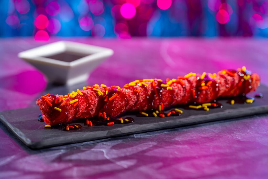 Red glazed dessert skewer coated in sugar with multicolored sprinkles on a dark serving slate.