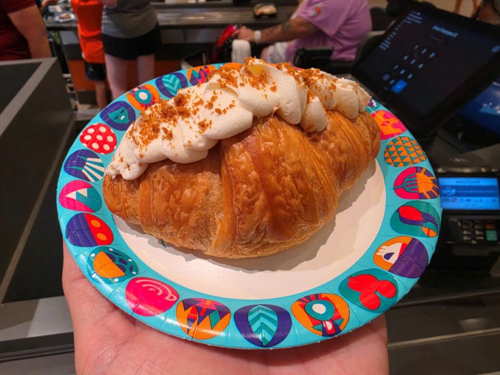 Cookie Butter Croissant topped with piped whipped cream and orange crumbs on a bright patterned plate at a cafe checkout area.