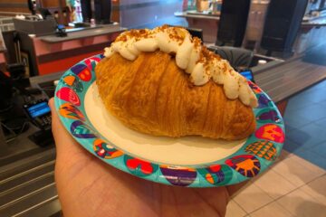 Cookie Butter croissant topped with whipped cream and crumbled bits on a colorful paper plate, held in a cafe.