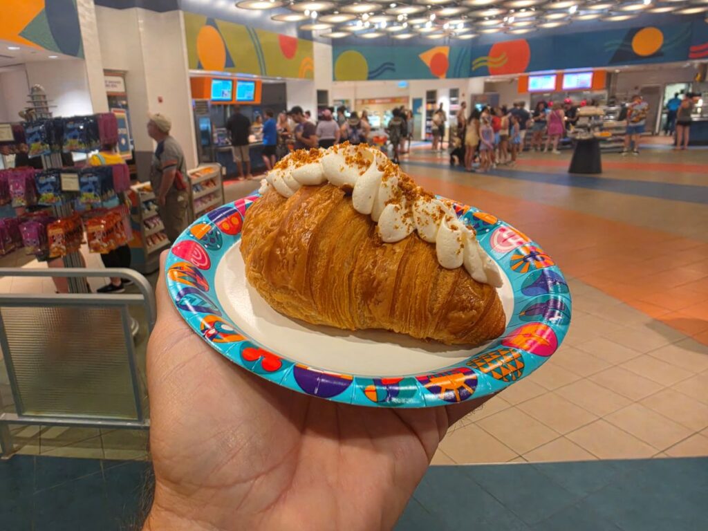 Flaky Cookie Butter Croissant topped with whipped cream on a colorful disposable plate, held up in a busy cafeteria/food court.