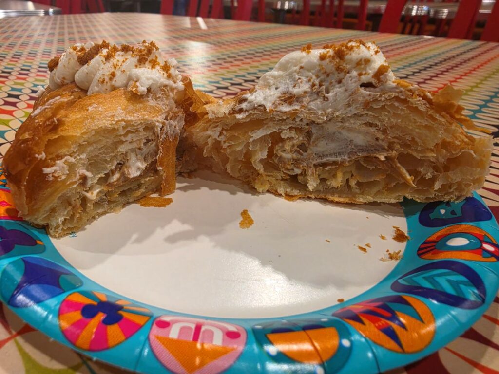 Flaky cream-filled pastry cut in half on a colorful paper plate, topped with whipped cream and crumbs.