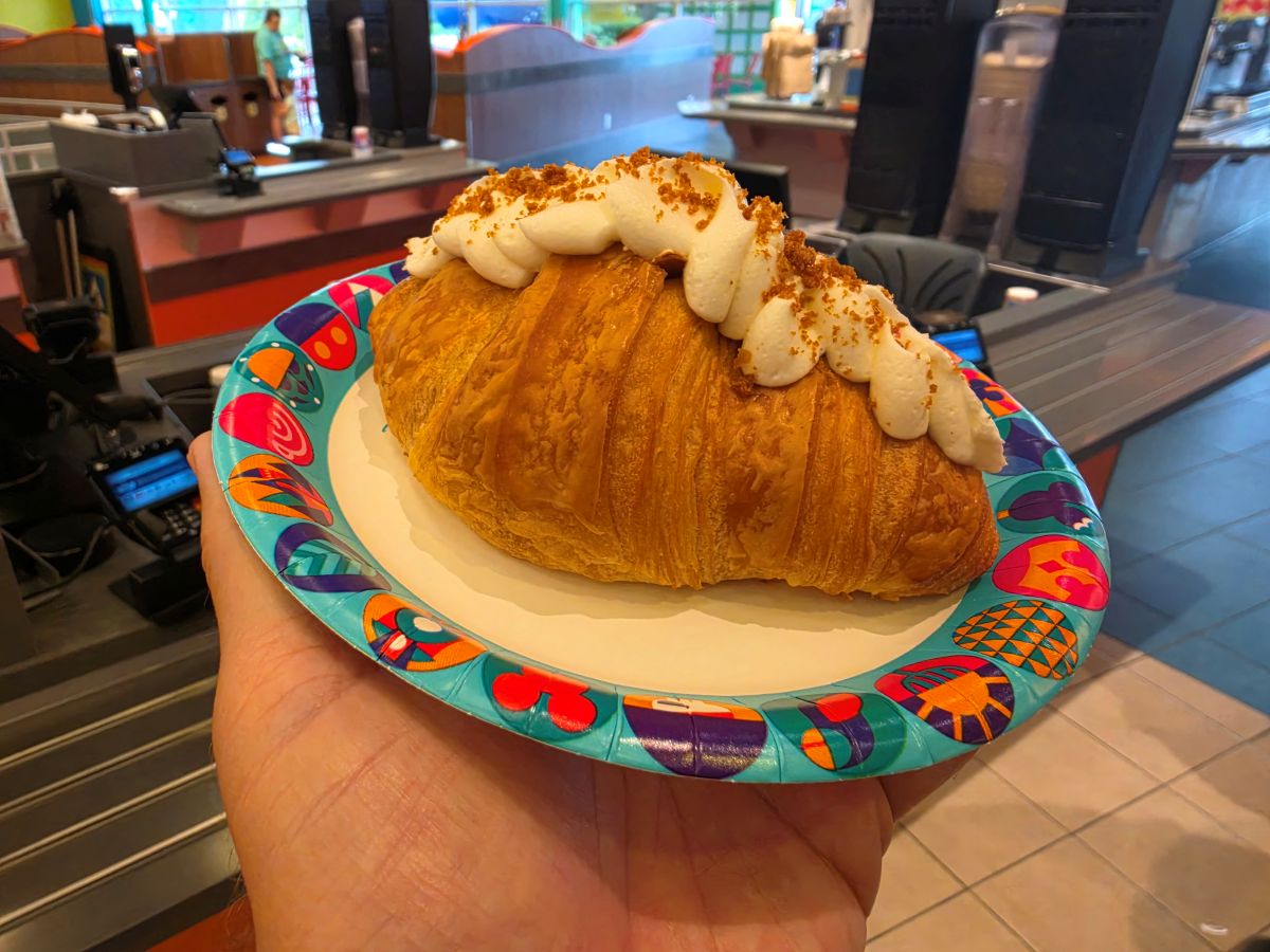 Cookie Butter croissant topped with whipped cream and crumbled bits on a colorful paper plate, held in a cafe.