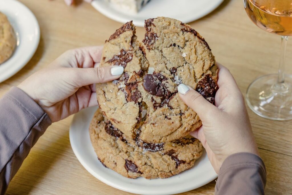 Hands break apart a large chocolate chip cookie over a white plate on a wooden table near a wine glass.