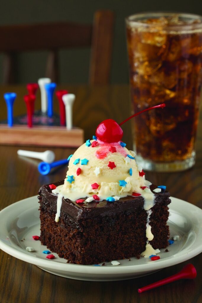 Chocolate cake square topped with a scoop of vanilla ice cream, a bright cherry, and red, white, and blue sprinkles; a glass of iced cola in the background.