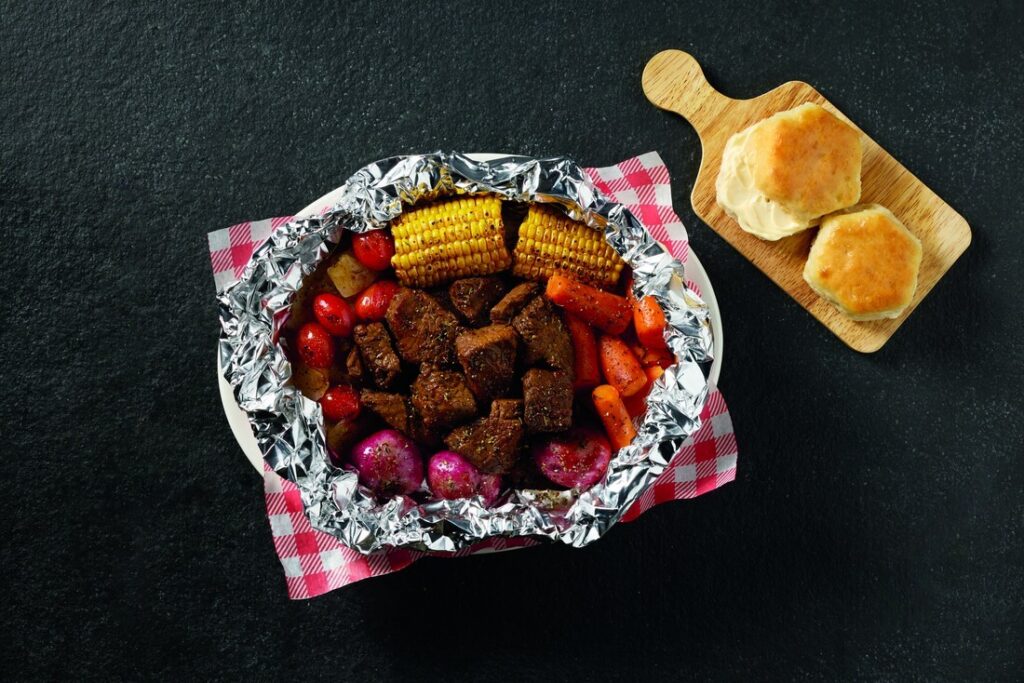 Beef and colorful roasted vegetables cooked in foil, served on a plate with corn, potatoes, carrots, and red onions atop a pink-checkered napkin. A wooden board with rolls sits nearby.