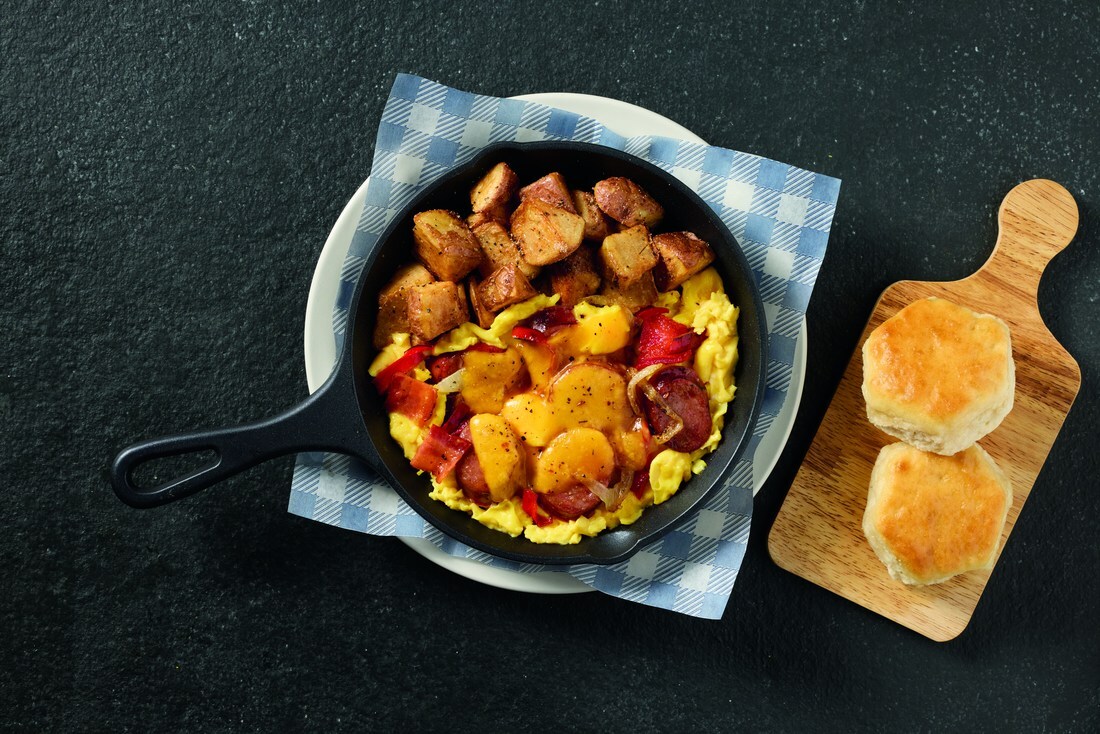 Skillet breakfast with roasted potatoes, red peppers, and scrambled eggs on a checkered napkin. Two buns sit on a wooden board nearby.