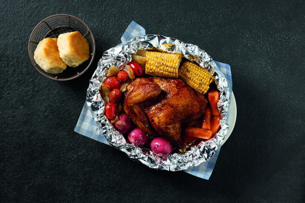 Top-down view of a roasted chicken with corn, potatoes, carrots, onions in a foil packet, served with a basket of bread rolls nearby.