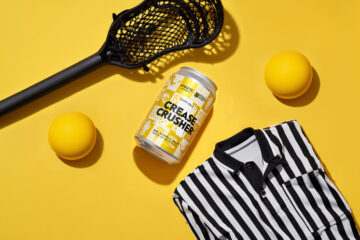 Lacrosse gear on a bright yellow background: stick head, two yellow balls, a striped polo shirt, and a can labeled 'Grease Crusher'.